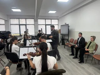 Un grup de muzicieni cu instrumente de coarde cântă într-o sală de clasă în fața a doi oameni așezați pe scaune.