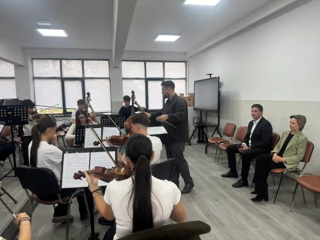 Un grup de muzicieni cu instrumente de coarde cântă într-o sală de clasă în fața a doi oameni așezați pe scaune.