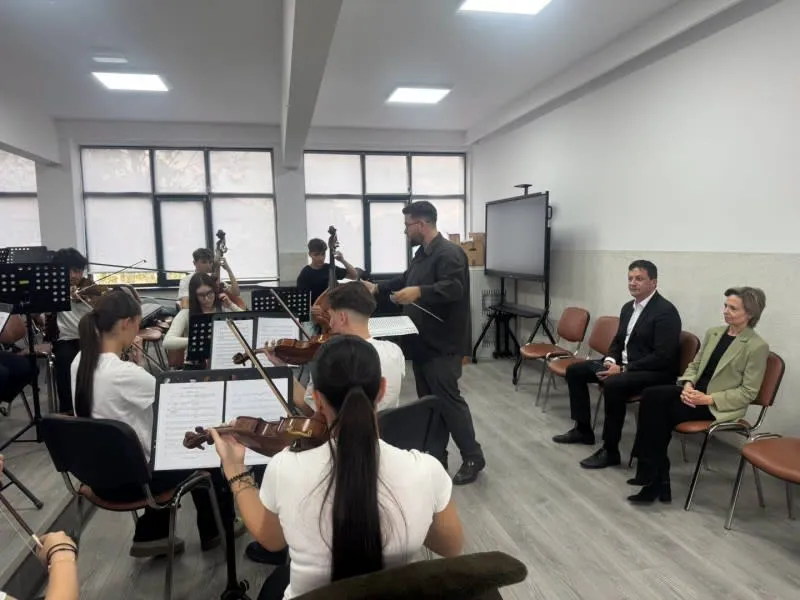 Un grup de muzicieni cu instrumente de coarde cântă într-o sală de clasă în fața a doi oameni așezați pe scaune.
