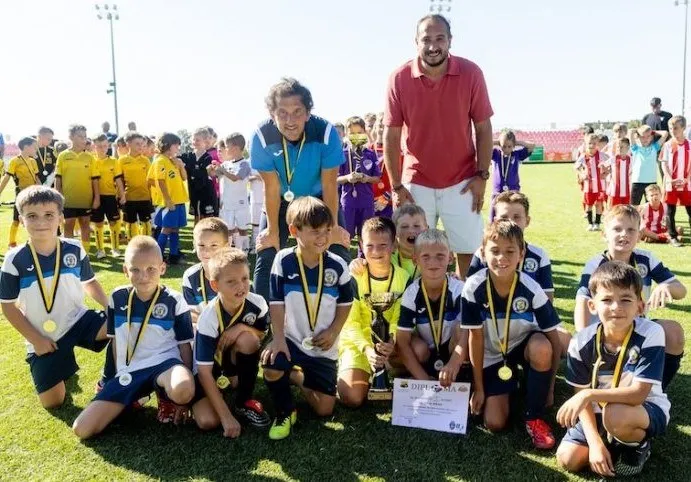 Un grup de copii și doi adulți stau pe un teren de fotbal verde, purtând medalii aurii și tricouri sportive colorate.