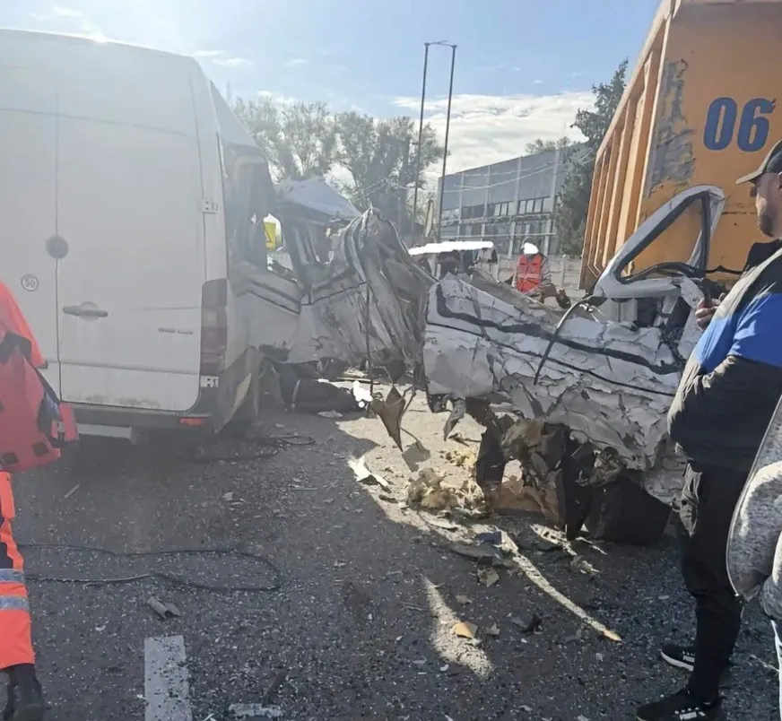 Un vehicul alb zdrobit stă pe carosabil lângă un microbuz alb și un camion galben, în prezența unor persoane.