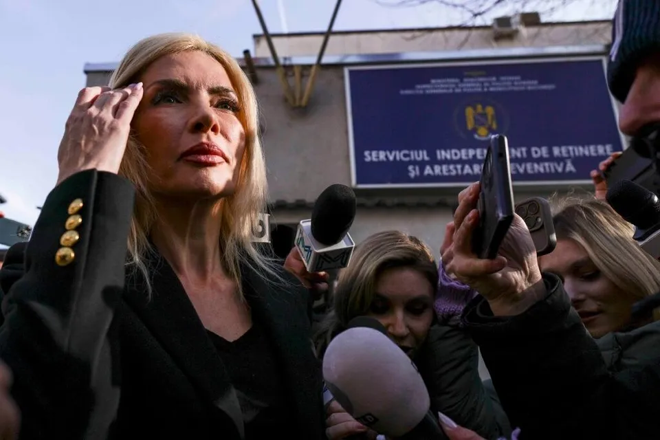 O femeie cu părul blond și haine negre este înconjurată de microfoane și telefoane mobile ridicate de persoane din jurul ei.