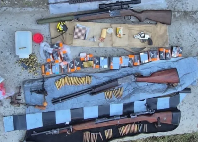 O imagine de sus a mai multor arme de foc, inclusiv puști și un pistol, alături de o cantitate mare de muniție, așezate pe o suprafață gri.