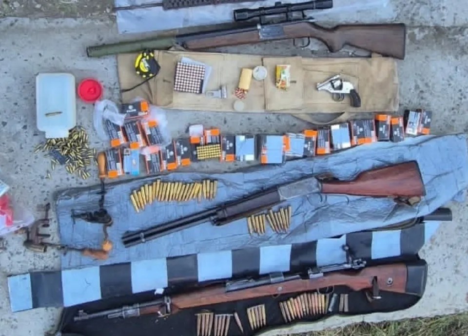 O imagine de sus a mai multor arme de foc, inclusiv puști și un pistol, alături de o cantitate mare de muniție, așezate pe o suprafață gri.