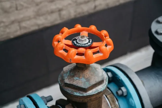 O roată de control portocalie este montată pe o supapă metalică oxidată, conectată la conducte de culoare albastră și neagră.