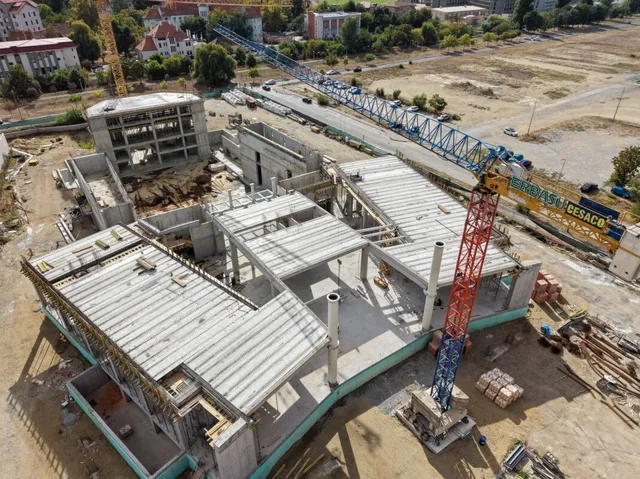 O vedere de sus prezintă un șantier cu structuri de beton gri, grinzi metalice și două macarale de construcții pe un teren de pământ.