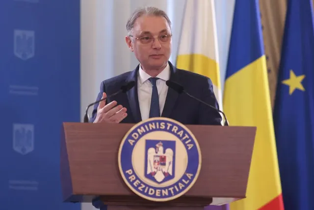 Un bărbat cu ochelari și costum închis stă în spatele unui podium de lemn pe care este afișat un emblema circulară colorată.