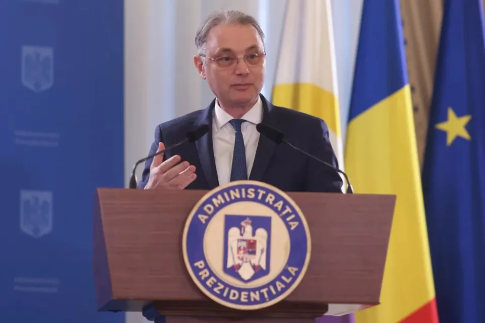Un bărbat cu ochelari și costum închis stă în spatele unui podium de lemn pe care este afișat un emblema circulară colorată.