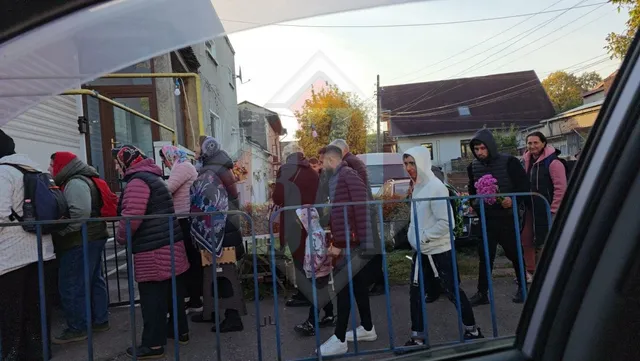O linie de oameni stă la coadă pe un drum strâns, fiind separați de o barieră metalică albastră.