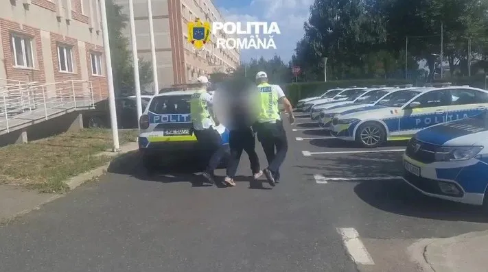 Trei polițiști în uniformă și veste reflectorizante conduc un bărbat cu fața blurată printre mai multe mașini de poliție parcate pe un teren asfaltat.