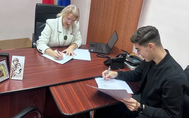 O femeie cu ochelari și un tânăr stau la două birouri separate, ambii scriind pe foi de hârtie albe cu pixuri.