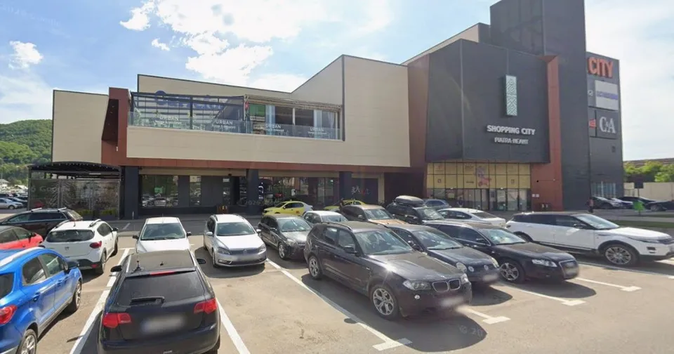 O clădire comercială cu fațadă în nuanțe de maro și cremă este situată în spatele unui parc de parcare cu diverse autoturisme colorate.