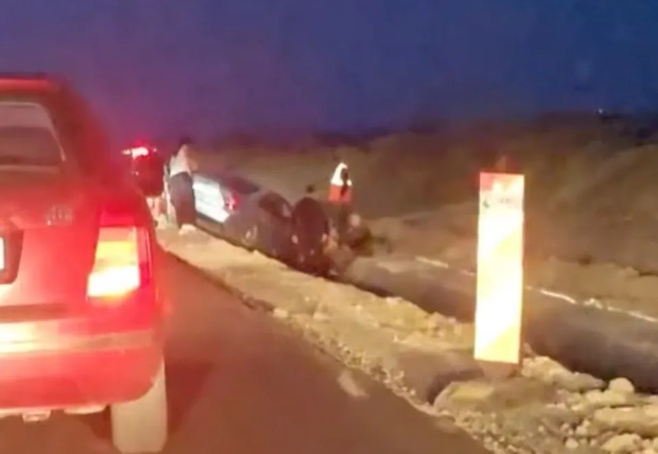 Un autoturism gri este poziționat într-un șanț de lângă un drum, lângă un vehicul roșu și un stâlp de semnalizare vertical.