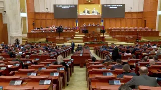 O sală de parlament cu rânduri de scaune din lemn ocupate de persoane și un podium central cu ecrane mari de proiecție.