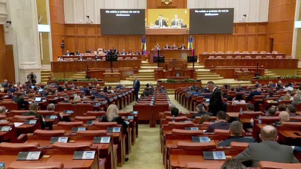 O sală de parlament cu rânduri de scaune din lemn ocupate de persoane și un podium central cu ecrane mari de proiecție.