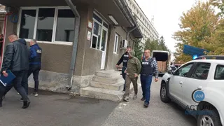 Cinci bărbați merg pe un trotuar lângă o clădire și o mașină albă, în timp ce un agent de poliție stă lângă un vehicul.