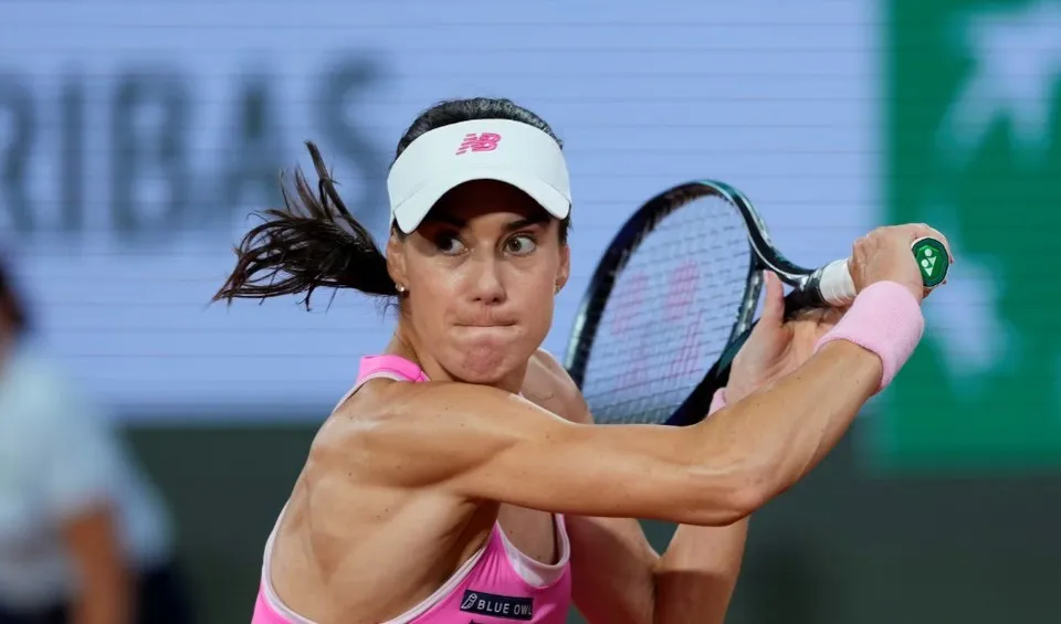 O femeie cu părul negru legat într-o coadă de cal lovește o minge cu o rachetă de tenis, purtând o vizieră albă și o ținută roz.
