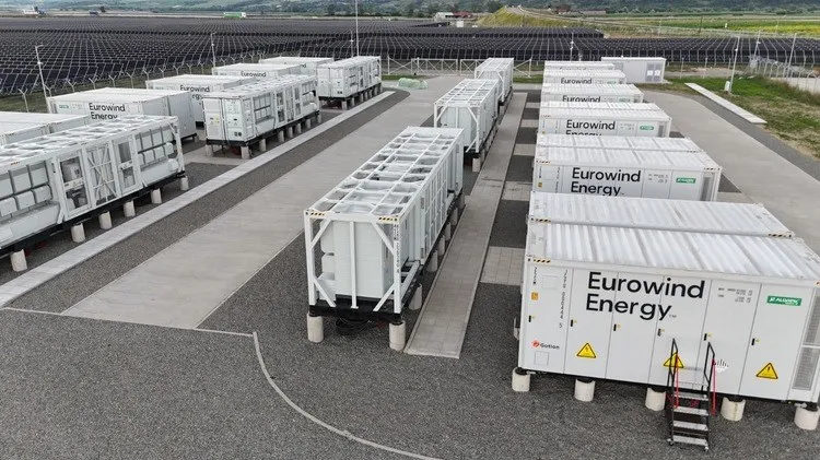 Mai multe containere albe cu inscripția Eurowind Energy sunt așezate pe platforme de beton într-o zonă industrială deschisă.