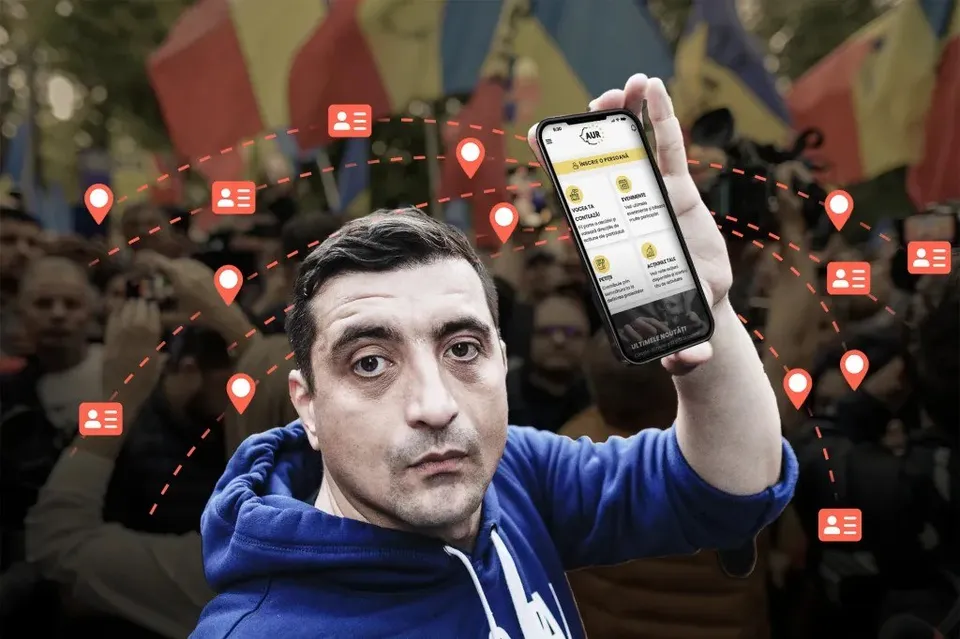 Un bărbat cu hanorac albastru ține un smartphone în mâna dreaptă în fața unei mulțimi de oameni și a unor steaguri colorate.