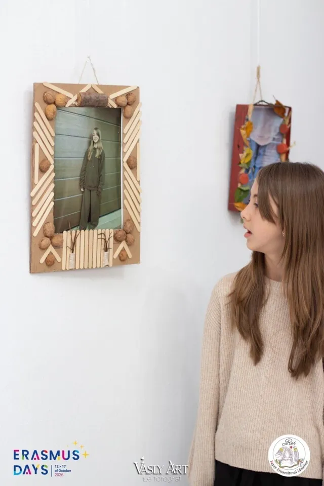 O tânără cu păr lung și deschis la culoră, îmbrăcată într-un pulover bej, se uită la o expunere de cadre foto decorate pe un perete alb.