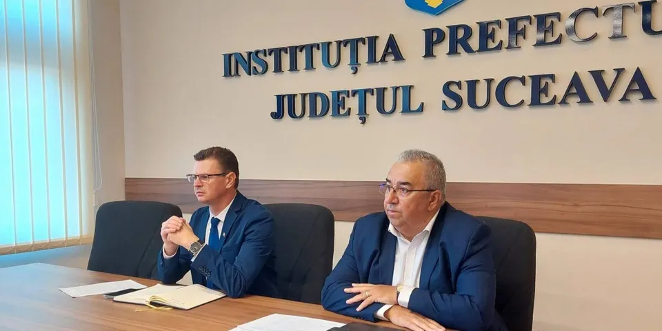 Un bărbat cu ochelari și bluza albă stă la o masă lângă un bărbat cu ochelari, cravată și sacou în fața unui perete cu text.