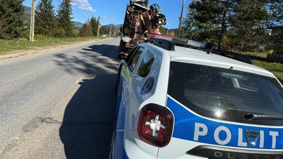 Un vehicul de poliție albastru și alb este parcat pe marginea unei drumuri asfaltate lângă o zonă cu vegetație și copaci.