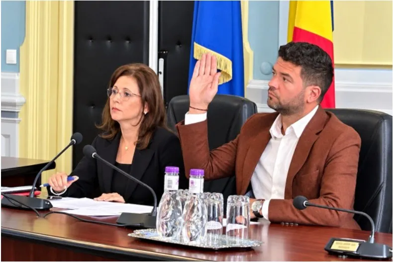 Un bărbat cu sacou maro ridică mâna dreaptă, stând lângă o femeie cu ochelari și sacou negru la o masă cu microfoane și sticle de apă.