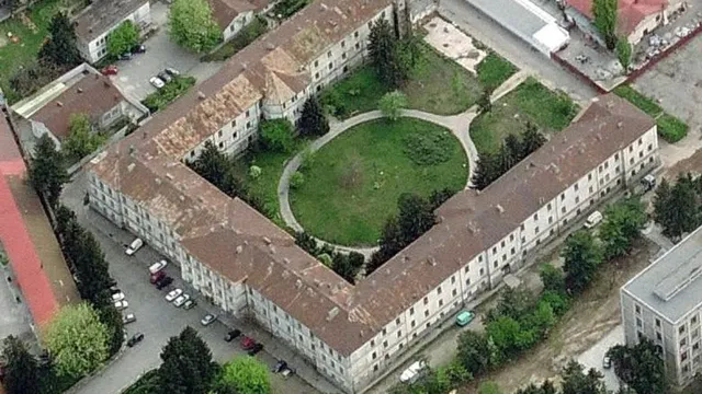 O vedere aeriană prezintă un complex de clădiri cu acoperișuri maronii dispuse în formă de U, înconjurând o curte verde cu alei.