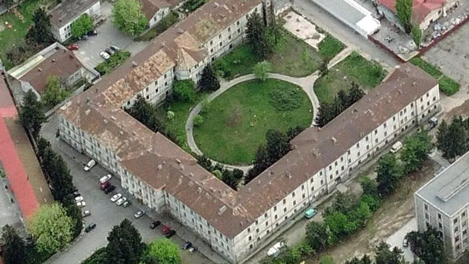 O vedere aeriană prezintă un complex de clădiri cu acoperișuri maronii dispuse în formă de U, înconjurând o curte verde cu alei.