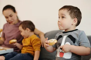 O femeie și doi copii mici stau pe o canapea, iar unul dintre copii ține în mână un produs de patiserie.