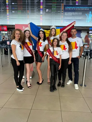 Un grup de șase copii și un adult stau în picioare în fața unui panou informativ mare într-un aeroport.