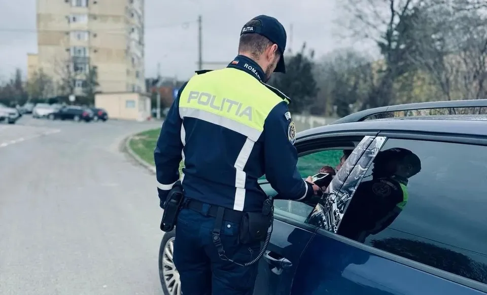 Un polițist în uniformă albastră și galbenă stă lângă fereastra deschisă a unei mașini gri pe un drum asfaltat.
