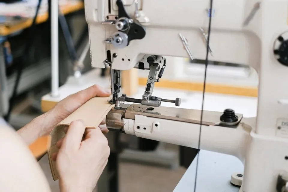 Mâinile unei persoane manipulează o bucată de material maro sub acul unei mașini de cusut industriale albe.