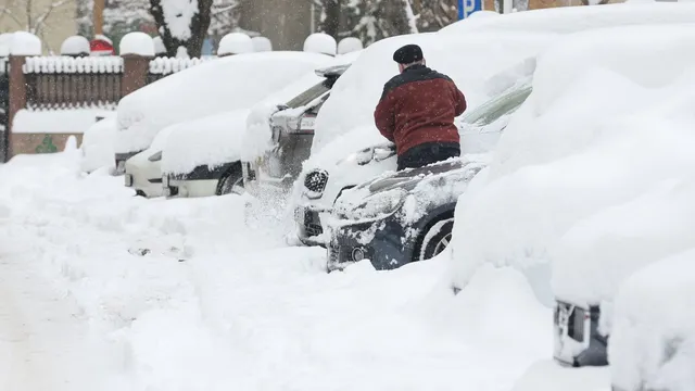 Un bărbat cu o jachetă roșie curăță zăpada de pe un autoturism parcat pe un drum acoperit de un strat gros de zăpadă.