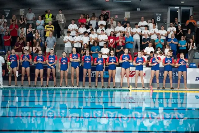 O echipă de water polo în echipament albastru și roșu stă în rând lângă piscina de la marginea bazinului, în fața unui public numeros.