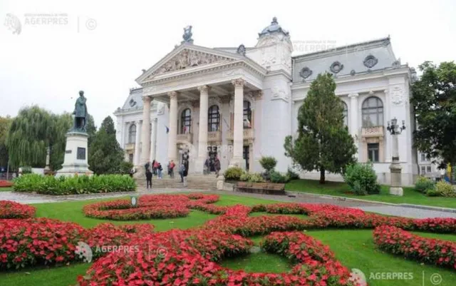 O clădire albă cu coloane și fronton se află în spatele unui gard de flori roșii și a unui monument cu o statuie.