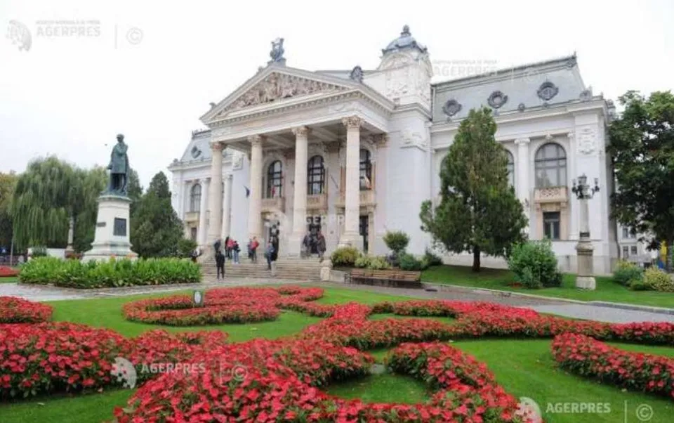 O clădire albă cu coloane și fronton se află în spatele unui gard de flori roșii și a unui monument cu o statuie.