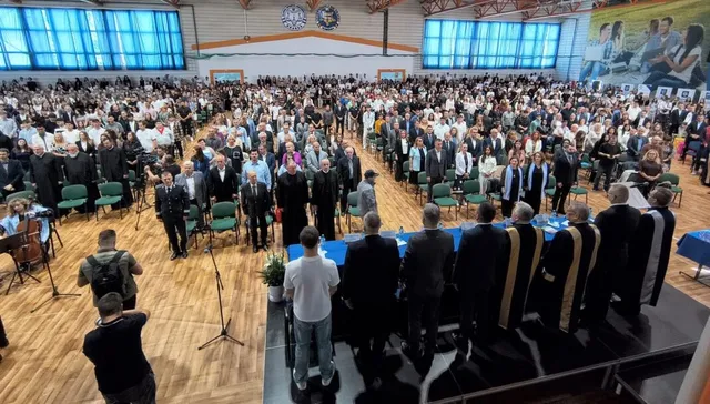 O mulțime de oameni stă în picioare într-o sală mare cu podea de lemn, în timp ce un grup de bărbați în costume negre stă pe scenă.