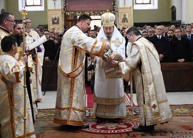 Câțiva bărbați în haine liturgice albe cu broderii aurii stau într-o biserică, în timp ce unul dintre ei ține un obiect metalic strălucitor.