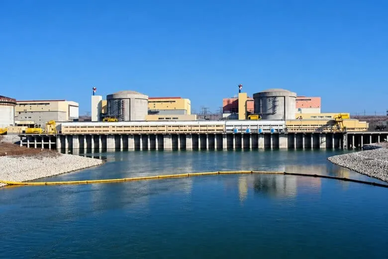 Două structuri cilindrice gri sunt situate lângă clădiri industriale lângă un corp de apă sub un cer senin.