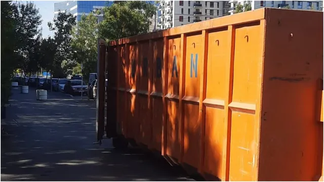 Un container metalic de culoare portocalie este amplasat pe un drum asfaltat lângă un grup de mașini parcate și copaci verzi.