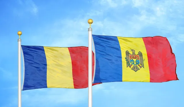 Două steaguri flutură pe catarge albe, în fața unui cer albastru cu nori subțiri.