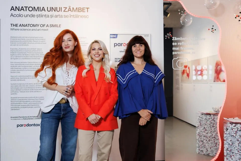 Trei femei stau în picioare în fața unui panou informativ alb, purtând bluze albe, un sacou roșu și o bluză albastră.
