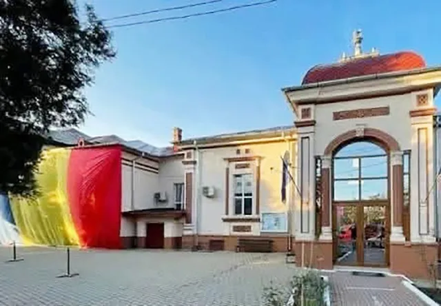 O clădire cu fațadă de culoare deschisă prezintă un pridvor înalt cu un arc mare și panouri de sticlă, alături de draperii colorate de dimensiuni mari.