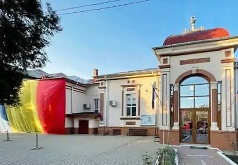 O clădire cu fațadă de culoare deschisă prezintă un pridvor înalt cu un arc mare și panouri de sticlă, alături de draperii colorate de dimensiuni mari.