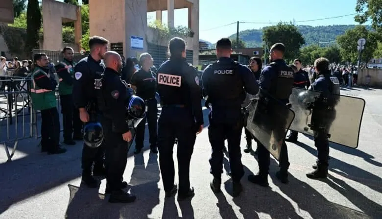 Mai mulți polițiști în uniforme închise și cu scrisul "POLICE" pe spate stau cu spatele, purtând echipament de protecție și scuturi transparente.