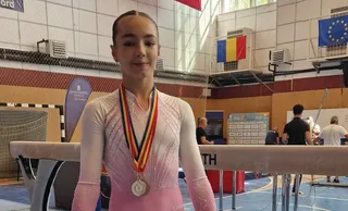 O tânără purtând o leotard roz cu sclipici și o medalie cu pangalică colorată stă în fața unui aparat de gimnastică într-o sală de sport.