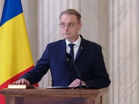 Un bărbat cu ochelari și costum închis la culoare stă în spatele unui pupitru de lemn lângă un steag tricolor.