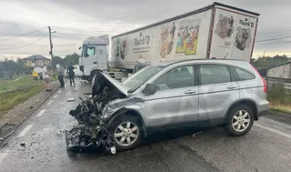 Un autoturism argintiu cu partea frontală distrusă este oprit pe un drum lângă un autotren alb cu reclame pe remorcă.