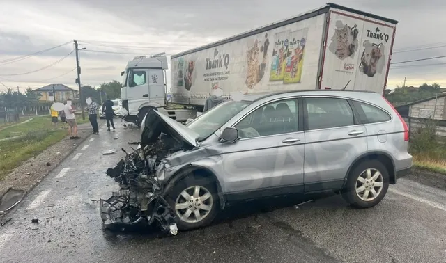 Un autoturism argintiu cu partea frontală distrusă este oprit pe un drum lângă un autotren alb cu reclame pe remorcă.